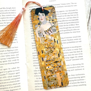 May include: A rectangular bookmark featuring a portrait of a woman in a gold and yellow dress, with a matching tassel. The artwork is reminiscent of Gustav Klimt's style, with intricate patterns and details. The bookmark is placed on an open book.