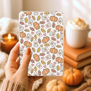 Peut inclure: Une coque de téléphone transparente avec un motif répété de citrouilles, de tasses à café, de cœurs et de feuilles d'automne en orange, rose, jaune et blanc. La coque est tenue dans une main, avec une bougie allumée et une tasse de café en arrière-plan.