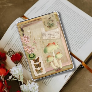 May include: A clear protective case with a vintage botanical design. The design features mushrooms, butterflies, and floral elements in pastel shades. The case is placed on an open book.