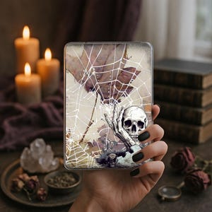 May include: A clear phone case featuring a spooky design. The case has a white spiderweb design over a purple leaf and a skull with a hand. The case is held in a hand with black nail polish.