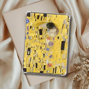 Könnte beinhalten: Eine Tablet-Hülle mit Gustav Klimts ikonischem Gemälde "Der Kuss". Die Hülle zeigt einen leuchtend gelben Hintergrund mit goldenen Akzenten und schwarzen rechteckigen Formen. Das Kunstwerk zeigt zwei Figuren, die sich umarmen, mit komplizierten Mustern und Farben.