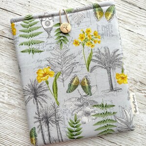 May include: A grey fabric book cover with a botanical design of yellow flowers, butterflies, ferns, and palm trees. It features a wooden button closure. A small tag reads "Bedish Baggies".