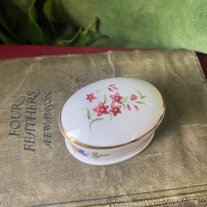 May include: A small, oval-shaped trinket box with a white porcelain base and a gold rim. The lid features a floral design with pink flowers and green leaves.