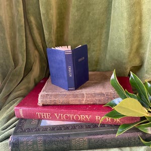 May include: A stack of vintage books with a small blue book on top. The blue book has the word "ENGLISH" printed on the spine. The books are on a green fabric surface.