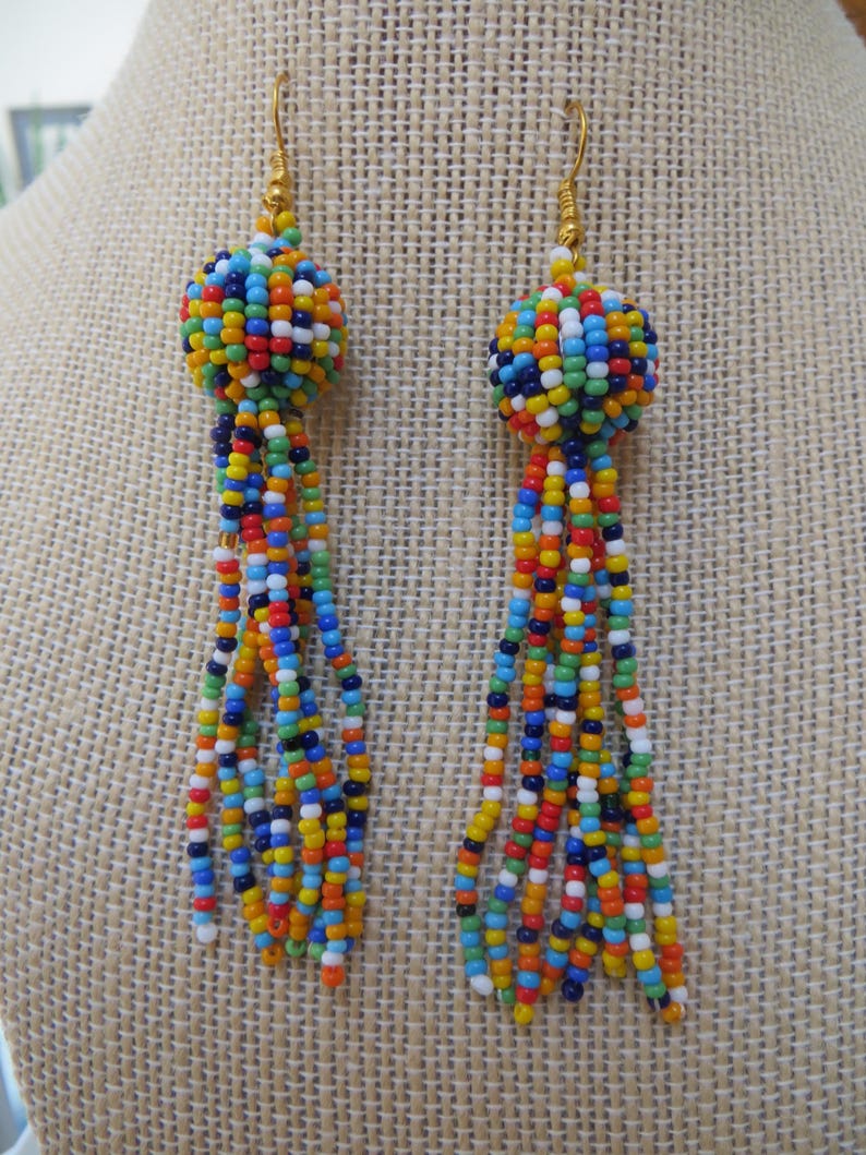 May include: A pair of colorful beaded earrings. Each earring features a round, beaded top in a mix of colors, with long beaded tassels in various colors. The earrings have gold-colored hooks.