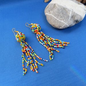 May include: A pair of beaded earrings with a ball-shaped top and long, dangling strands. The earrings feature a vibrant mix of colorful beads in shades of blue, orange, yellow, green, and red. The earrings are set against a bright blue background.