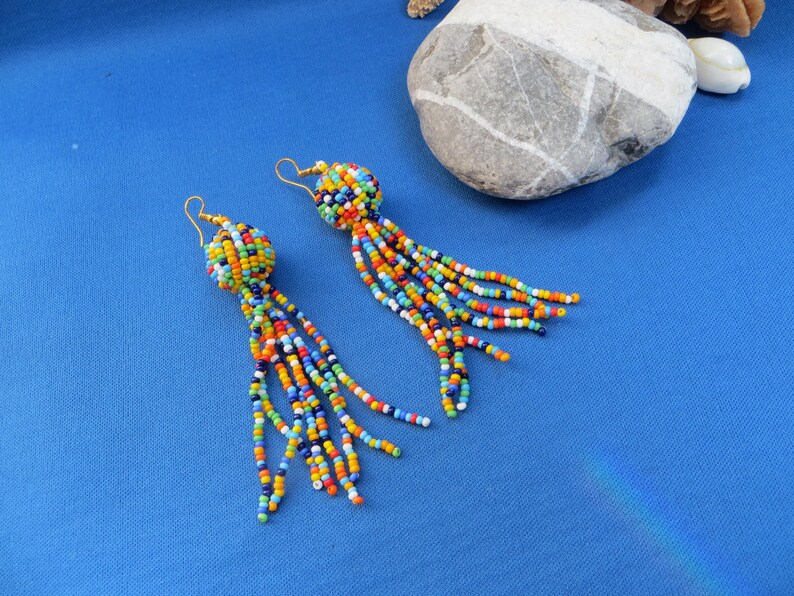 May include: A pair of beaded earrings with a ball-shaped top and long, dangling strands. The earrings feature a vibrant mix of colorful beads in shades of blue, orange, yellow, green, and red. The earrings are set against a bright blue background.