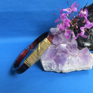May include: A dark red and gold bracelet with a woven gold band. The bracelet is displayed on a blue surface next to a cluster of purple amethyst crystals and purple flowers. A dark sculpture is in the background.