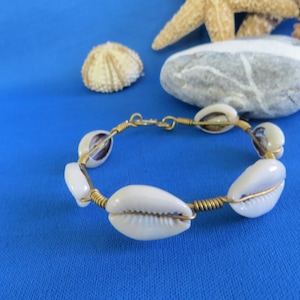 May include: A bracelet featuring white cowrie shells connected by gold-toned wire. The shells are arranged in a circular pattern, with the wire wrapped around each shell to secure them in place. The design includes a simple metal clasp closure.