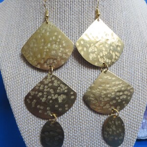 May include: A pair of gold-colored earrings with a hammered texture. Each earring features a fan-shaped top, a smaller fan shape below, and an oval dangle. The earrings hang from gold-colored ear wires.