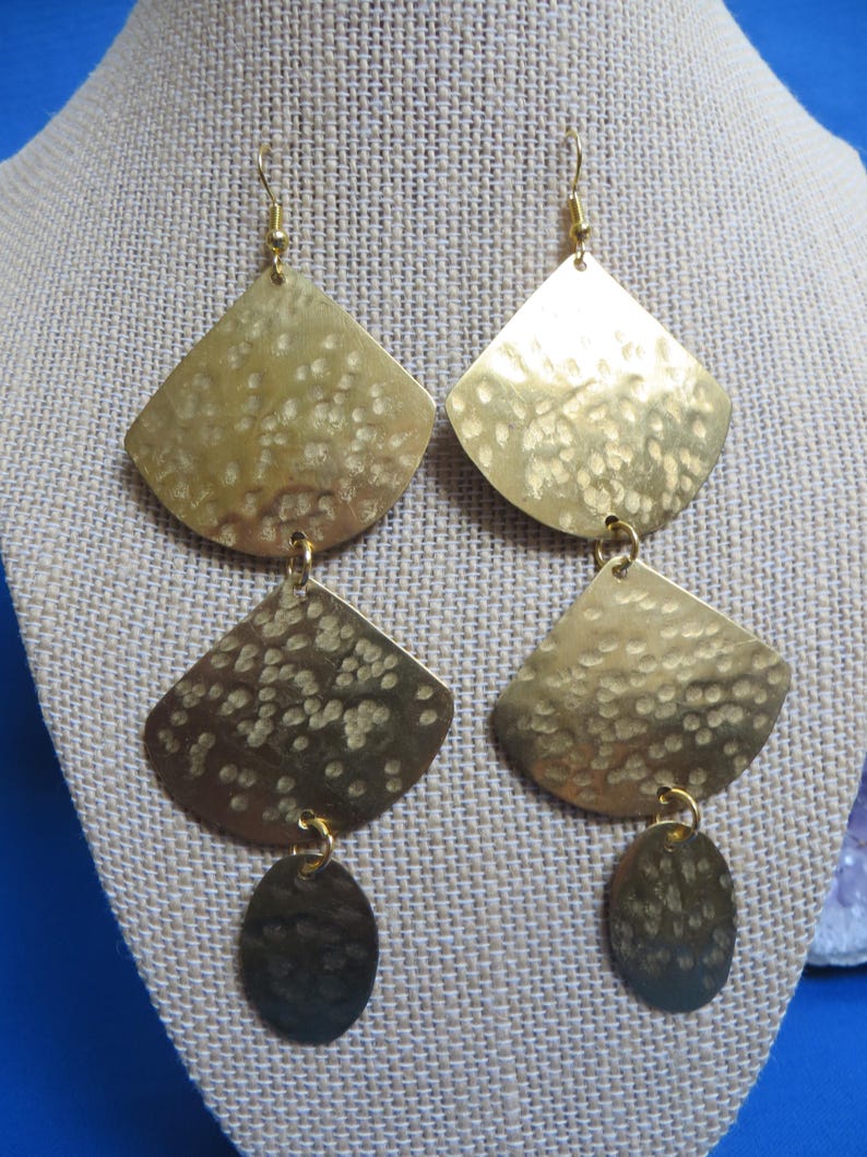 May include: A pair of gold-colored earrings with a hammered texture. Each earring features a fan-shaped top, a smaller fan shape below, and an oval dangle. The earrings hang from gold-colored ear wires.