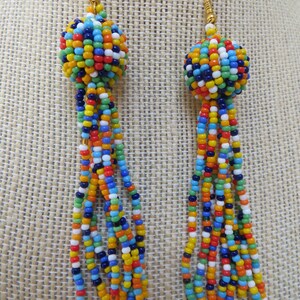 May include: A pair of colorful beaded earrings. Each earring features a spherical cluster of small beads in various colors, including blue, orange, yellow, and white. Long strands of matching beads dangle from the sphere, creating a tassel effect. The earrings have gold-colored hooks.