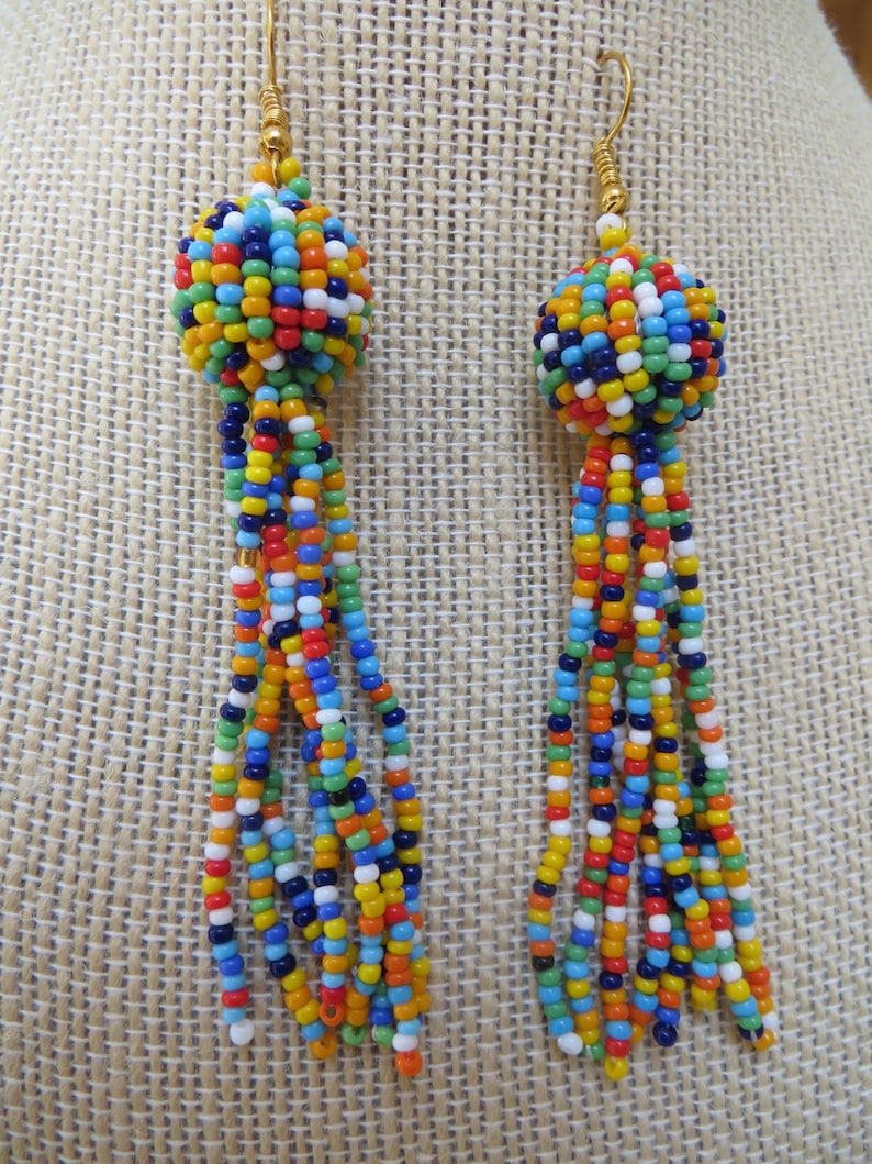 May include: A pair of colorful beaded earrings. Each earring features a spherical cluster of small beads in various colors, including blue, orange, yellow, and white. Long strands of matching beads dangle from the sphere, creating a tassel effect. The earrings have gold-colored hooks.