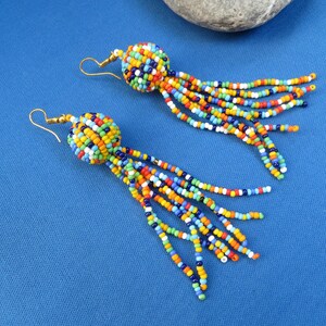 May include: A pair of colorful beaded earrings with gold-colored hooks. Each earring features a round, beaded top in a mix of colors, with long beaded strands in various colors hanging down. The earrings are set against a blue background.