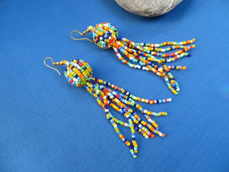 May include: A pair of colorful beaded earrings with gold-colored hooks. Each earring features a round, beaded top in a mix of colors, with long beaded strands in various colors hanging down. The earrings are set against a blue background.