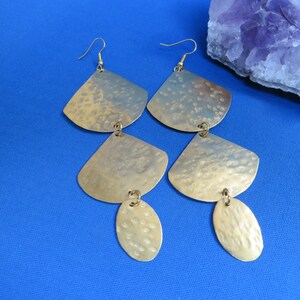 May include: Gold-toned earrings with a hammered texture. Each earring features a fan-shaped top piece, a smaller, oval-shaped bottom piece, and a hook for pierced ears. The earrings are displayed on a blue surface.