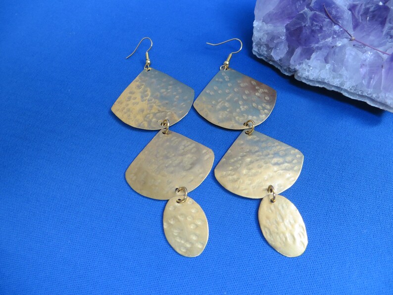 May include: Gold-toned earrings with a hammered texture. Each earring features a fan-shaped top piece, a smaller, oval-shaped bottom piece, and a hook for pierced ears. The earrings are displayed on a blue surface.