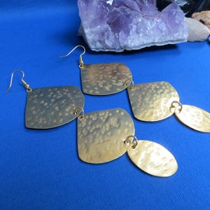 May include: Gold-toned, hammered metal earrings with a unique design. Each earring features a square shape connected to an oval shape, all suspended from gold-colored ear wires. The earrings have a textured surface and are displayed on a blue background.