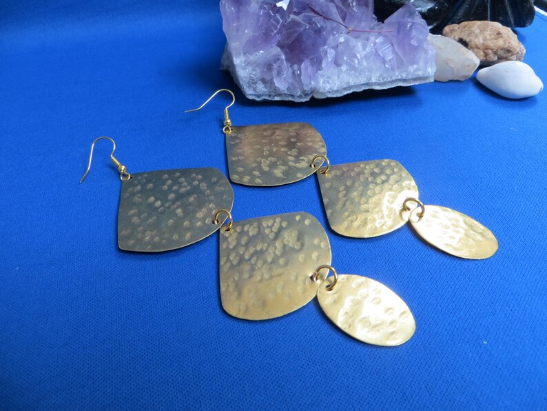 May include: Gold-toned, hammered metal earrings with a unique design. Each earring features a square shape connected to an oval shape, all suspended from gold-colored ear wires. The earrings have a textured surface and are displayed on a blue background.