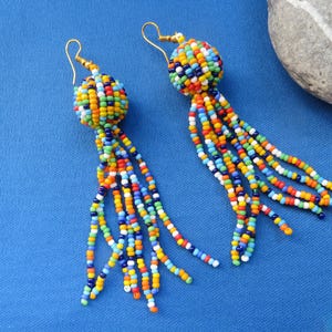 May include: A pair of handmade beaded earrings. Each earring features a round, multicolored bead cluster with dangling strands of colorful beads. The earrings have gold-colored ear wires and are set against a blue background.