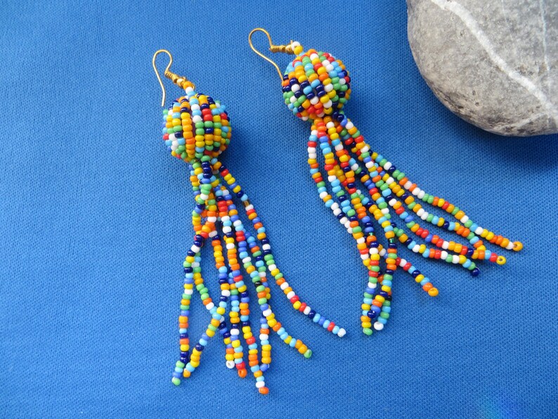 May include: A pair of handmade beaded earrings. Each earring features a round, multicolored bead cluster with dangling strands of colorful beads. The earrings have gold-colored ear wires and are set against a blue background.