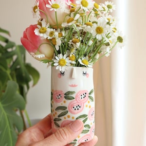 Puede incluir: Un jarrón de cerámica blanco con diseño de rostro, lleno de tulipanes rosas y margaritas blancas. El jarrón tiene un patrón floral rosa con hojas verdes. Las flores se sostienen en una mano.