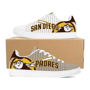 May include: White canvas low-top sneakers with a brown and yellow baseball-themed design. The design features a cartoon character swinging a bat with the words "San Diego Padres" on the shoe.