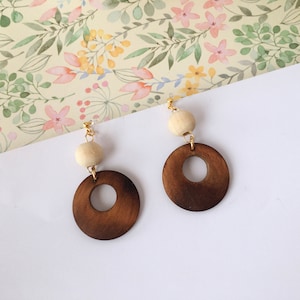 May include: A pair of wooden earrings with a brown circle and a white bead. The earrings are on a white background with a floral pattern.