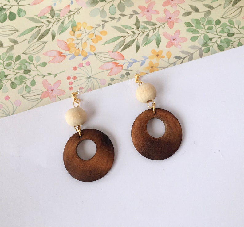 May include: A pair of wooden earrings with a brown circle and a white bead. The earrings are on a white background with a floral pattern.