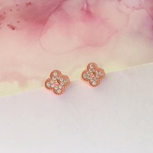 May include: A pair of rose gold earrings with a four-leaf clover design. Each earring is adorned with small, clear cubic zirconia stones.