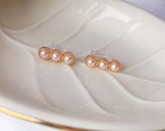May include: A pair of clip-on earrings featuring three small, pale pink pearls set in a gold-colored, beaded frame. The earrings are displayed on a white, leaf-shaped dish.