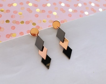 Könnte beinhalten: Ein Paar geometrischer Hängeohrringe. Jeder Ohrring hat einen kleinen, runden, roségoldenen Stecker, der mit drei absteigenden Diamantformen in Schwarz und Roségold verbunden ist. Die Ohrringe werden auf einer weißen Oberfläche mit rosa Hintergrund präsentiert.
