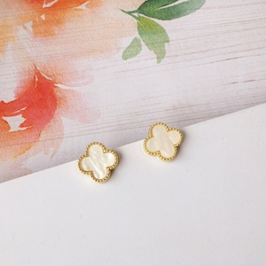 May include: Two gold-colored clover-shaped earrings with a white center. The earrings have a textured gold border and are set against a white and floral background.