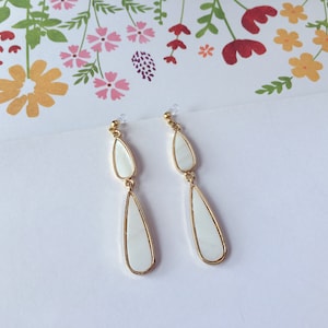 May include: A pair of gold dangle earrings with white teardrop-shaped stones. The earrings are made of metal and have a simple design.