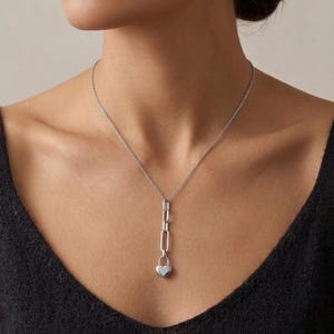 May include: A silver necklace featuring a chain design with a heart-shaped pendant. The necklace is worn on a person wearing a black top. The pendant is a small, polished heart. The necklace is a delicate piece of jewelry.