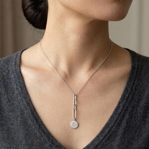 Peut inclure: Collier argenté composé d'une fine chaîne et d'un pendentif vertical avec des formes liées et un petit disque rond. Le collier est porté sur un haut gris.