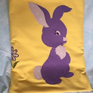 May include: Yellow pillowcase featuring a purple bunny applique with a white fluffy tail and chest. The bunny has a light pink inner ear and a flower applique in the lower left corner. The pillowcase is rectangular.