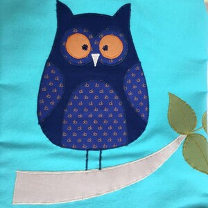 May include: A blue owl with orange eyes and a white beak perched on a branch with green leaves. The owl has a pattern of small brown and blue squares on its body. The background is turquoise.