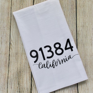 May include: White kitchen towel with the black text "91384 California". The towel is folded and placed on a light-colored wooden surface. The text is in a bold, sans-serif font with the word "California" in a cursive font.