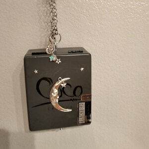 May include: A gray wooden box with a silver crescent moon charm hanging from it. The box has the word "Coco" written on it and a sticker that says "Blackbird" on the side. The box is decorated with silver stars.
