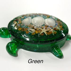 May include: A green turtle-shaped trinket box with a clear, glossy finish. The lid features seashells, small stones, and starfish embedded in a clear resin. The word "Green" is printed at the bottom.