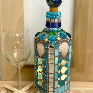 May include: A decorative bottle with a mosaic design featuring shades of blue, turquoise, and silver, adorned with seashells. The bottle has a matching mosaic stopper and is next to a wine glass and a starfish.