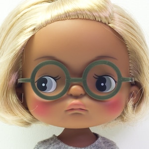 May include: A doll with blonde hair and brown skin wearing round green glasses and a grey shirt.