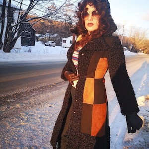 May include: A woman wearing a long, brown, fuzzy coat with a patchwork design. The coat has a brown, black, and orange color scheme. She is wearing sunglasses and has long, curly hair. The woman is standing on a snowy road.