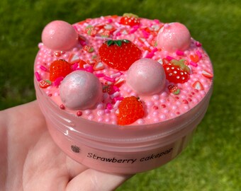 May include: Pink slime with white and pink sprinkles, red and white candy hearts, and red and white strawberry slices. There are also large white and pink balls on top of the slime. The slime is in a clear plastic container with a pink lid. The container has a label that says "Strawberry cakepop".