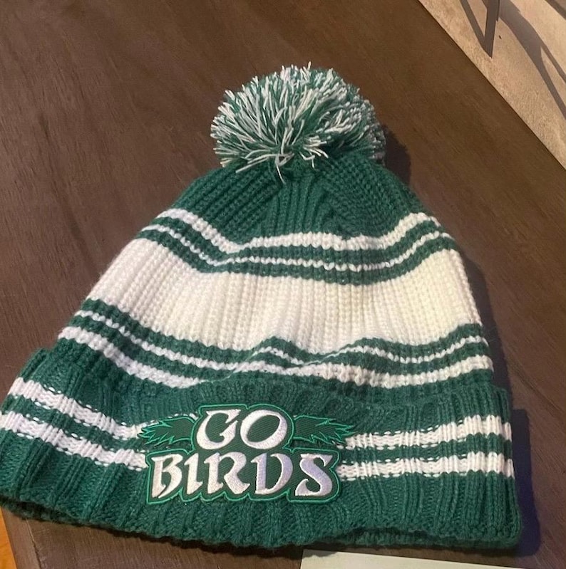 May include: Green and white striped knit beanie with a white pom pom on top. The beanie has a green embroidered patch that says "GO BIRDS".