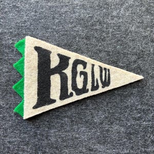 Puede incluir: Un banderín de fieltro color crema con las letras "KGLW" en negro, con un borde en zigzag verde. El banderín tiene forma triangular y descansa sobre una superficie gris. Las letras están en una fuente audaz y estilizada.