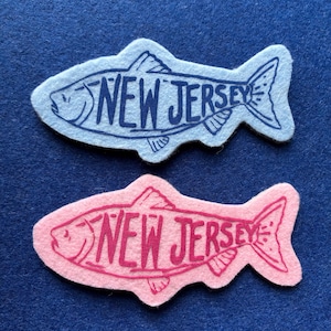 Könnte beinhalten: Zwei filzige Aufnäher in Fischform, einer hellblau mit dunkelblauer Tinte und einer hellrosa mit dunkelrosa Tinte. Beide zeigen eine handgezeichnete Umrisslinie eines Fisches mit dem Text NEW JERSEY, der mittig auf den Körper gedruckt ist.