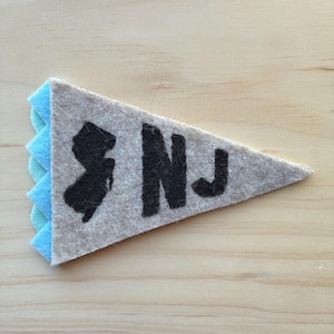 Puede incluir: Un banderín de fieltro gris en forma de triángulo, con el contorno negro de Nueva Jersey y las letras "NJ". Tiene un borde festoneado azul claro.