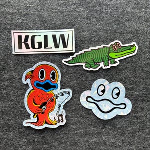 May include: A collection of four holographic stickers. One rectangular sticker with the letters "KGLW". Other stickers include a green alligator, a red cartoon character fishing, and a white cloud with eyes.